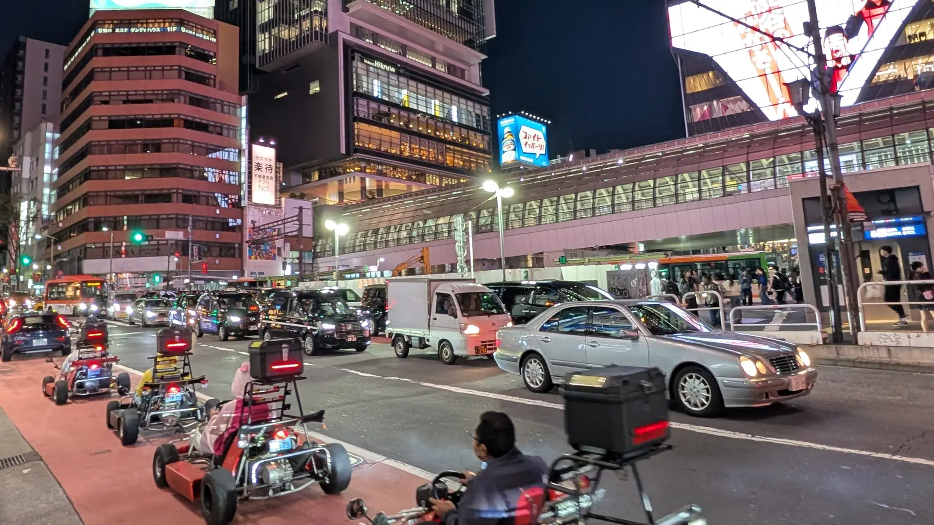 Street-legal go-karts driving through a neon-lit Tokyo crossing at night, surrounded by city traffic and illuminated shopfronts